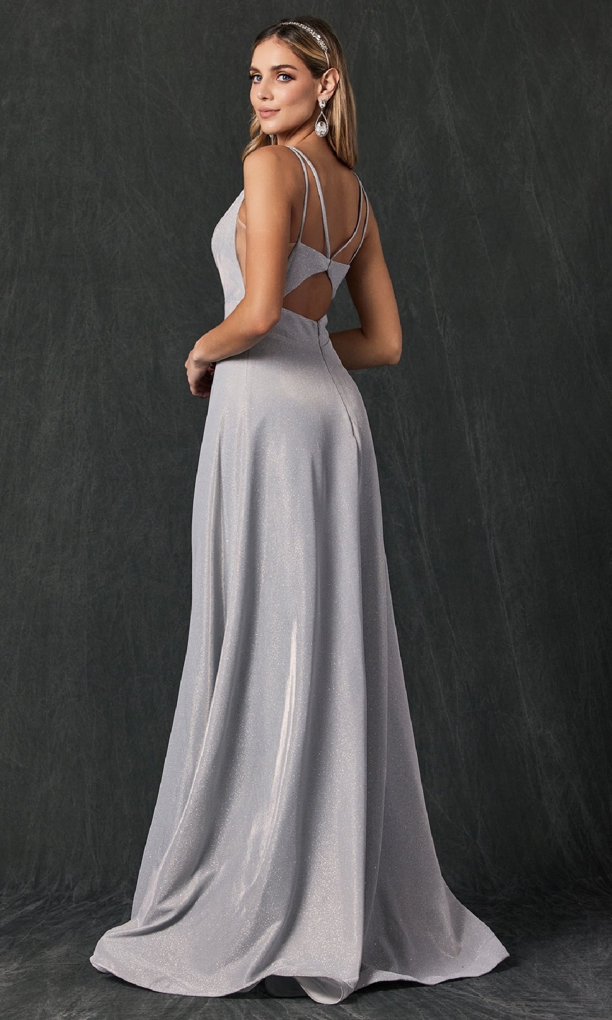 A woman wears the Juliet 208 long prom dress in silver, featuring a sleeveless design and open back, as she stands slightly turned and looks over her shoulder against a dark, textured backdrop.